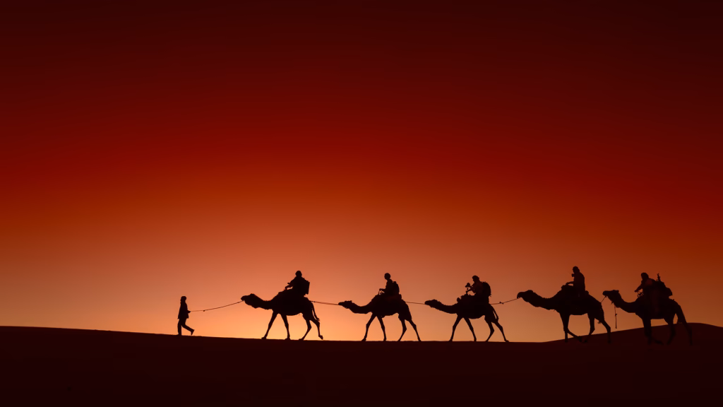 Silhouette of a camel caravan crossing a dune at sunset under a red sky